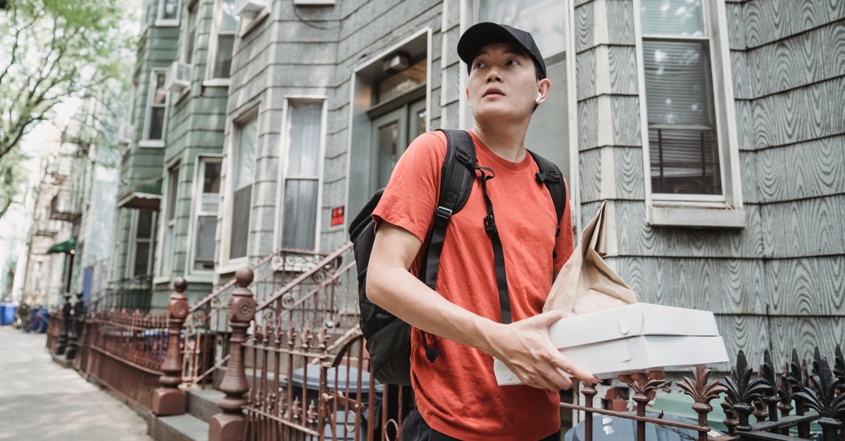 A delivery worker in a red shirt and cap holding packages outside a residential building.